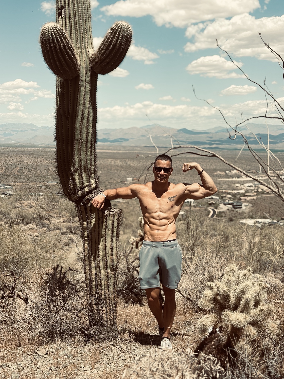 Casey Barnard standing beside a tall cactus in Arizona, flexing his arm with mountains behind him.