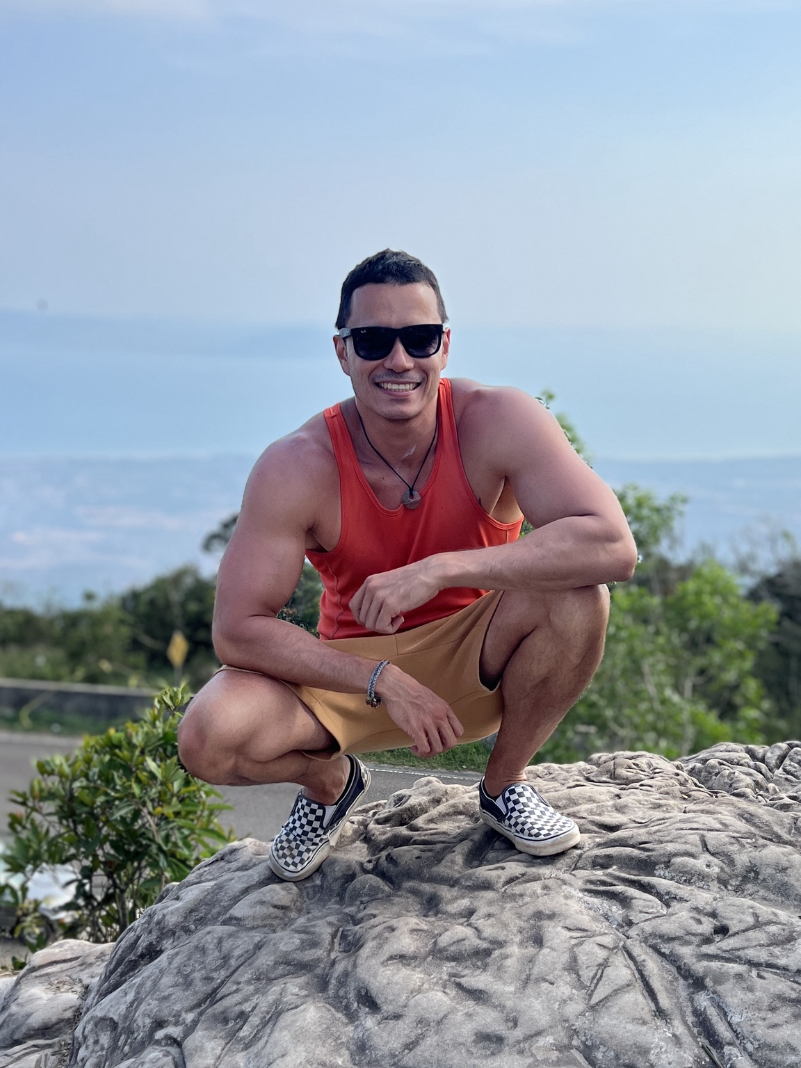 Casey Barnard smiling in a red singlet and sunglasses, crouching on a rock outdoors.